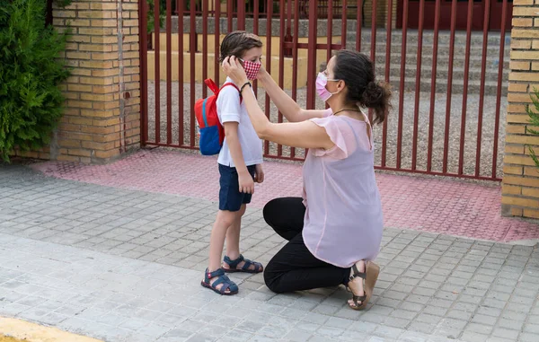 Okulun kapısında okul çocuğuna yüz maskesi takan bir kadın. Covid19 salgını sırasında okuldaki yeni normalin konsepti.