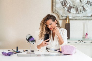 Kafasında havlu olan beyaz bir kadın makyaj yaparken kendi portresini çekiyor..