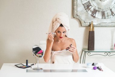 Kafasında havlu olan beyaz bir kadın aynanın önünde makyaj yapıyor..