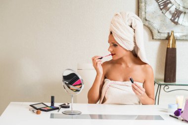 Kafasında havluyla beyaz bir kadın aynanın önünde dudaklarını boyuyor..