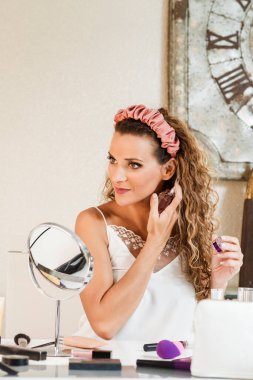 Genç sarışın beyaz kadın makyaj yapıyor ve aynanın önünde parfüm sıkıyor..