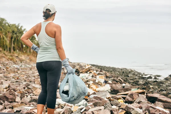 Женщина в перчатках держит пластиковый мусор на грязном побережье — стоковое фото