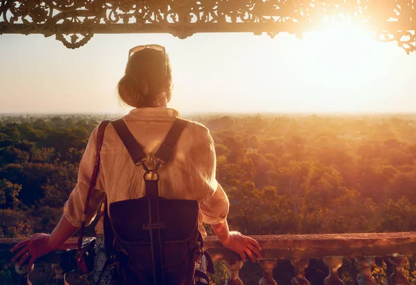 Vrouw toerist met een rugzak staat en kijkt naar de zonsondergang boven het bos op een oud houten balkon. — Stockfoto