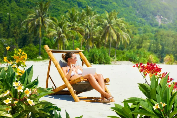 Donna felice con computer portatile in sedia di bambù che lavora a distanza sulla spiaggia tropicale — Foto Stock