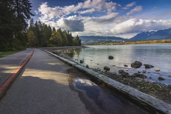 Winding coastal Stanley Park Seawall Path on a sunny day with dramatic cloudscape over Vancouver Harbor and Lions Gate Bridge in the distance, Stanley Park, Vancouver, British Columbia