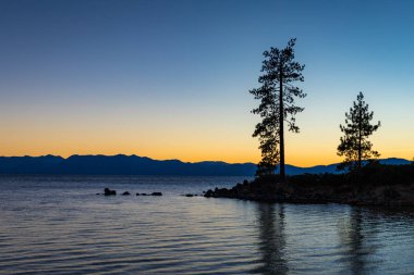 Günbatımında sakin su, ağaçlar ve arka plan, Lake Tahoe, Carson City, Nevada dağlarında siluetleri ile açık gökyüzü kum Harbor