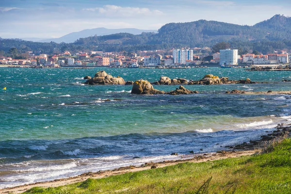 Coastal rocks and wind waves with Vilanova de Arousa seafront at background