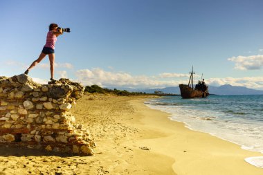 Tourism vacation and travel. Woman tourist on beach taking photo with camera, shipwreck in the background clipart