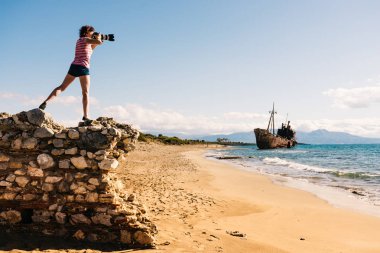 Tourism vacation and travel. Woman tourist on beach taking photo with camera, shipwreck in the background clipart