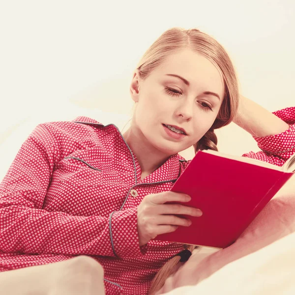女の子は読書本に横たわっていた マットレスの上で家でリラックスした若いブロンドの女性が赤い点線のパジャマを着ています トーンフィルターされた画像 — ストック写真