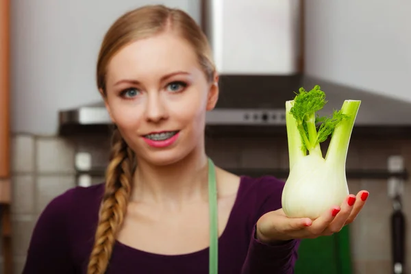 Woman in kitchen holding green fresh raw fennel bulb vegetable. Young housewife cooking. Healthy eating, vegetarian food, dieting and people concept.