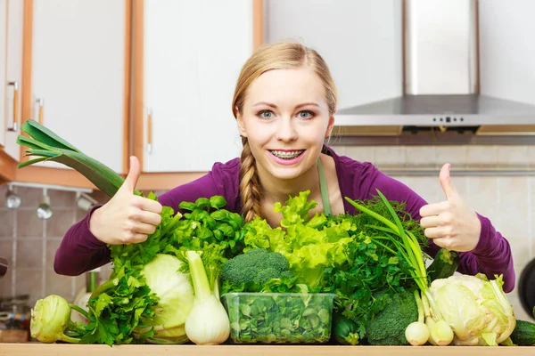 Woman in kitchen with many green leafy vegetables making thumb up hand sign gesture. Young housewife adding to her diet foods high in chlorophyll. Healthy eating, cooking, vegan food concept.