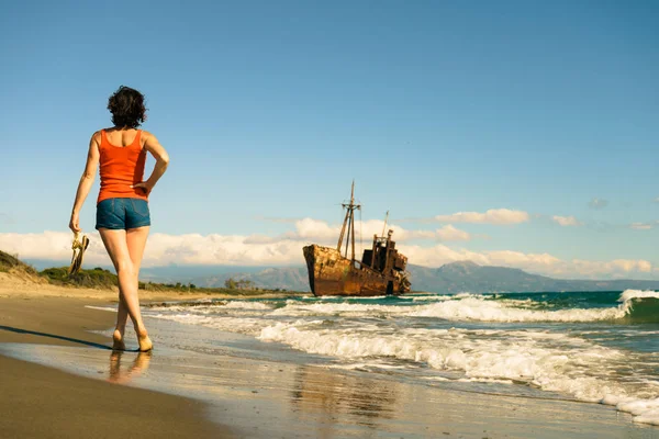 旅の自由 夏休みを楽しむビーチを歩く熟女観光客の女性 古い放棄された難破船 バックグラウンドで難破船 — ストック写真
