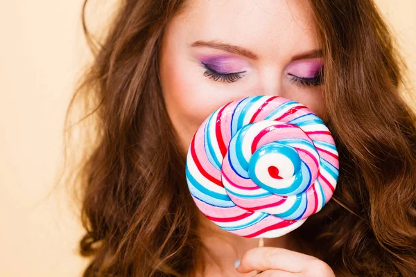 Woman attractive girl colorful eyes make up holding big lollipop candy in hand, covering her face. Sweet food and enjoying concept. Studio shot on bright