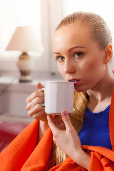 暖かい毛布の下でソファの上に寝そべっている若い女性は冷たい天候の間に彼女の余暇時間を楽しんでマグカップから紅茶 コーヒーまたは別の熱い飲み物を飲む — ストック写真