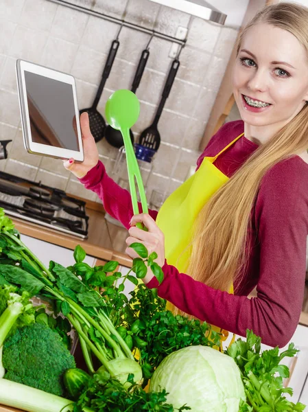 台所の若い女性はテーブルの上に多くの緑の野菜を持っています 何かを調理し インターネットでレシピを検索することについて考えるタブレットを保持 — ストック写真