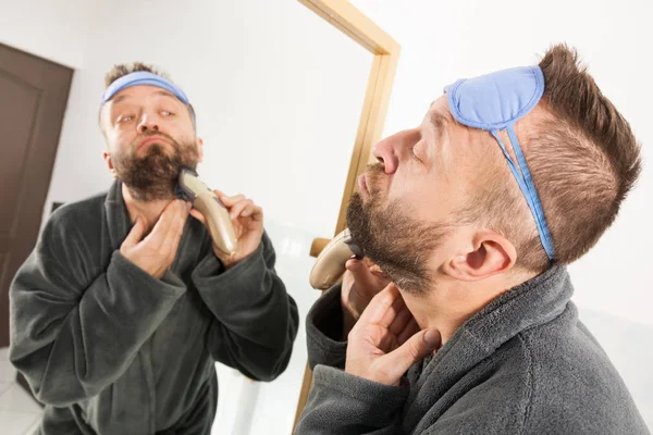 ひげを生やした男は鏡の中で自分自身を見て 電動タイマーカミソリを使ってひげを剃った 男は顔の毛の世話をし 眠いアイマスクを着て — ストック写真