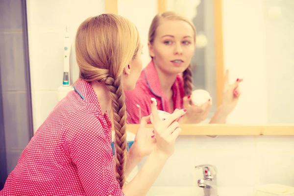 若いブロンドの女性がバスルームミラーを見て 顔に保湿スキンクリームを適用します 乾燥した色白層の保湿剤の世話をする女の子 スキンケア治療 — ストック写真