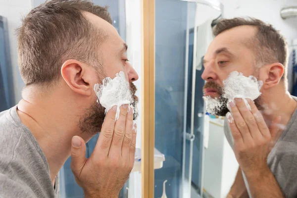 Man preparing his facial hair before trimming his beard, applying shaving cream foam mousse. Male beauty treatment concept.