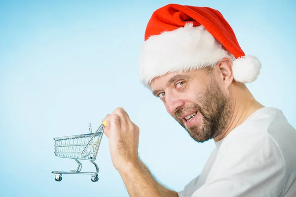 青い空のショッピングバスケットカートを保持男はサンタのクラスの帽子を身に着けている クリスマス 季節販売 ボクシングの日のコンセプト — ストック写真