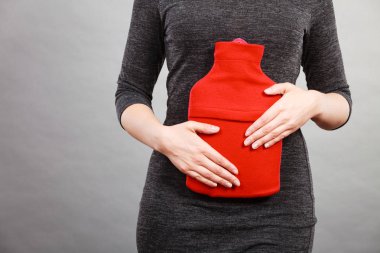 Unrecognizable woman having strong stomach ache. Female suffer on belly pain, holding hot red water bottle on abdomen. Health care, remedy for pains concept clipart