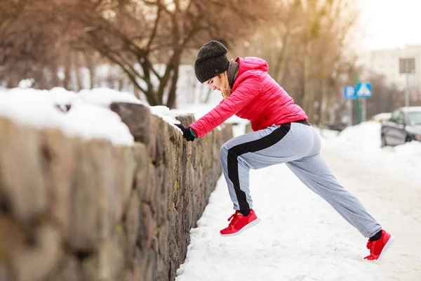 Outdoor sport exercises, sporty outfit ideas. Woman wearing warm sportswear training exercising stretching legs outside during winter.