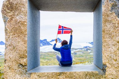 Tourist woman holds norwegian flag, enjoying mountains scenery from stone sculpture at Mefjellet viewpoint. National route 55 Sognefjellet, Norway. Jotunheimen National Park. clipart