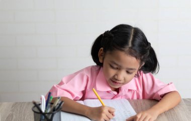 Küçük kız mutlu ev ödevi yapıyor. Çocuklar kullanım sarı bir kalem bir defter ahşap masanın üzerine yazıyor. Odak alan sığ derinliği seçin.