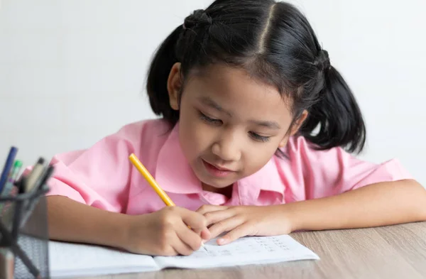 Yakın yukarı küçük kız mutlu ev ödevi yapıyor. Çocuklar kullanım sarı bir kalem bir defter ahşap masanın üzerine yazıyor. Odak alan sığ derinliği seçin.