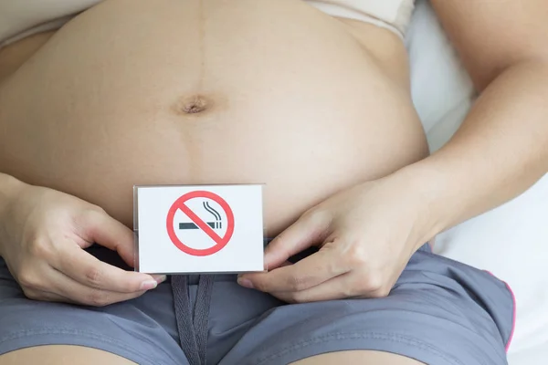 Pregnant woman holding label with smoking ban in bedroom background, unhealthy concept.