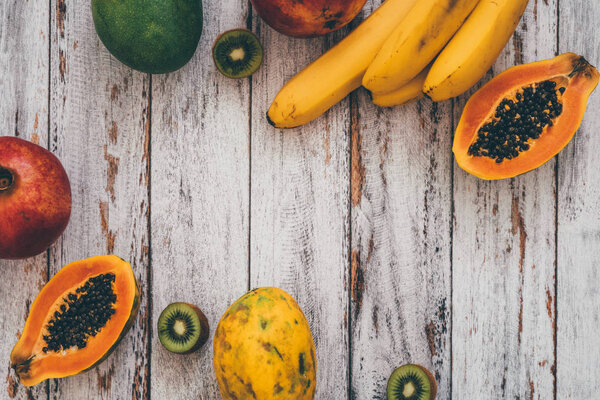 Вид сверху на целые манго, бананы, гранаты, лаймы, скользящие папайи и киви на деревянном фоне
