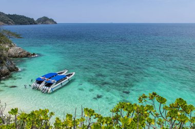 İki sürat teknesi denize park etmiş. Tekne beyaz görünüyor ve mavi bir çatıya sahiptir. Berrak mavi deniz, ışık yansıması, Küçük dalgalar.