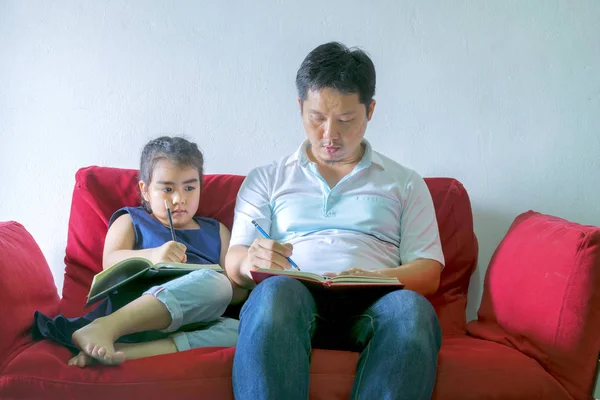 Father and daughter are writing books.Sitting on a red sofa,It is a good learning practice and a good family relationship.