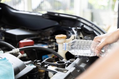 Arabasının radyatörünü dolduran bir kadın. Arabanın açık kaputunun yanında duruyor, bir elinde bir şişe su tutuyor ve diğeriyle birlikte radyatöre döküyor..