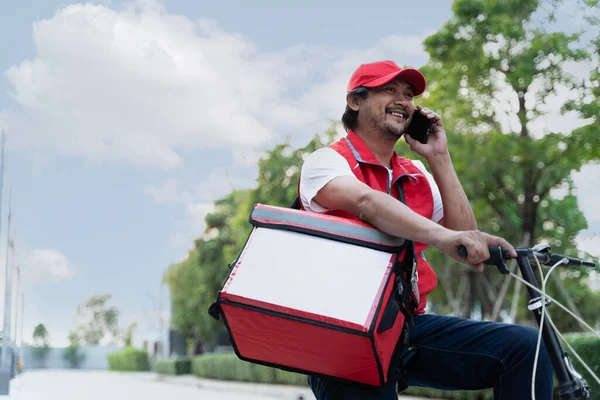 A delivery person riding a bicycle while using a smartphone. The person is wearing a helmet and a uniform, and is holding the phone to their ear with one hand while steering the bike with the other.