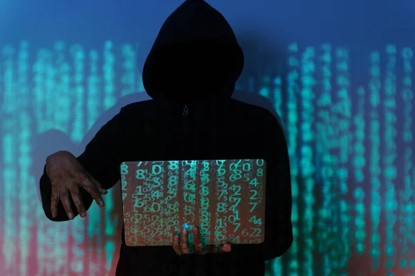 A hacker is sitting in a dark room, hunched over a laptop computer. The hacker is wearing a hoodie and sunglasses, and their face is hidden in the shadows. The laptop screen is filled with code.