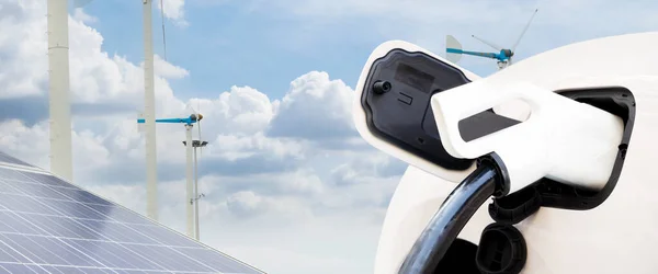 An electric car is plugged into a charging station. The charging station is powered by solar panels and a wind turbine. The car is parked in a parking lot. The sun is shining and the wind is blowing.