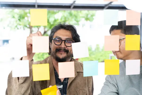 Business people reading adhesive notes on glass wall in office. Business people meeting at office and using post notes to share ideas, brainstorming ideas, sticky notes on glass wall.