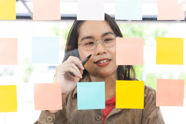 Confident business woman wearing glasses. Write your ideas or tasks on sticky paper on the glass wall. Female team leader, executive manager, holding a computer tablet Planning an event project