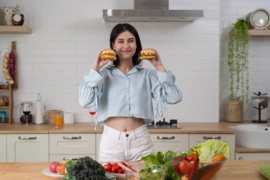Mutfakta elinde taze sebzeler ve meyvelerle iki hamburger tutan mutlu kadın, sağlıklı yaşam tarzı, beslenme, gıda seçimi, denge, sağlık, organik malzemeler, yemek pişirme, tazelik konsepti..
