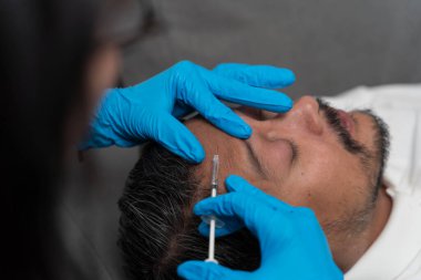 Uzanıp alnına mavi eldiven giyen profesyonel bir doktor tarafından estetik enjeksiyon alan adama yakın çekim, güzellik tedavisi kavramı, cilt bakımı, cilt bakımı, dermatoloji, tıbbi prosedür ve klinik.