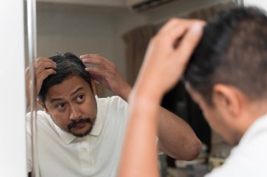 Orta yaşlı bir adamın aynaya bakıp gri saçlarını endişeyle incelemesi, yaşlanmayı, saç dökülmesini, sağlık sorunlarını, kendine bakımı ve günlük hayatta kişisel görünüm bilincini sembolize etmesi..