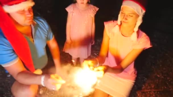 Счастливая семья с ребенком в рождественских шляпах горит бенгальские огни в руках — стоковое видео