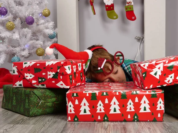 Niño en Santa sombrero y gafas de Navidad se duermen detrás de los regalos de Navidad —  Fotos de Stock