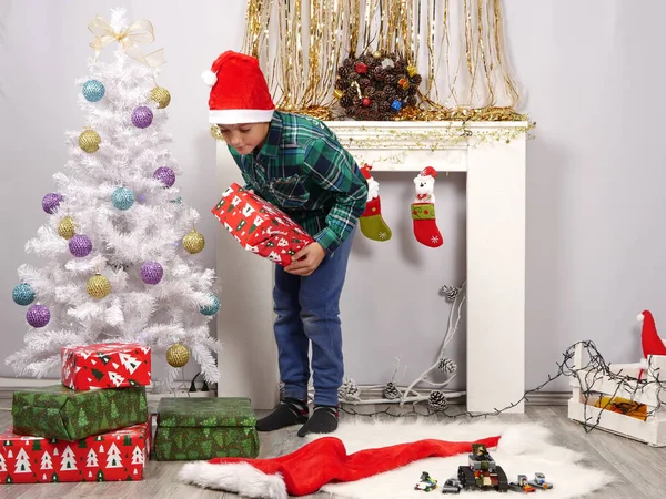Fiú Santa kalap teszi bemutatja a karácsonyfa közelében — Stock Fotó