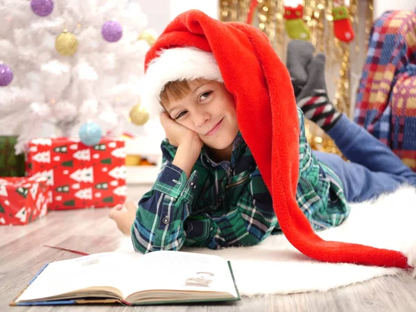 Fiú hosszú karácsony kalapban Olvasson egy könyvet a karácsonyfa közelében — Stock Fotó