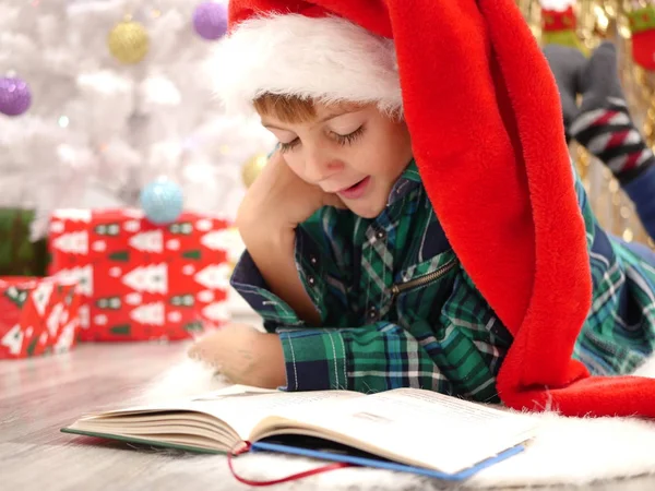 Fiú hosszú karácsony kalapban olvasó egy könyv és ásít a karácsonyfa közelében — Stock Fotó