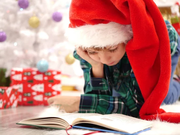 Fiú hosszú karácsony kalapban Olvasson egy könyvet a karácsonyfa közelében — Stock Fotó