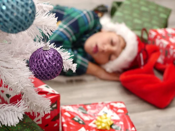 Fiú alszik a földön mögött a karácsonyfa karácsony kalapban — Stock Fotó