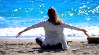 Young woman sitting cross-leged near the sea and streching in windy weather